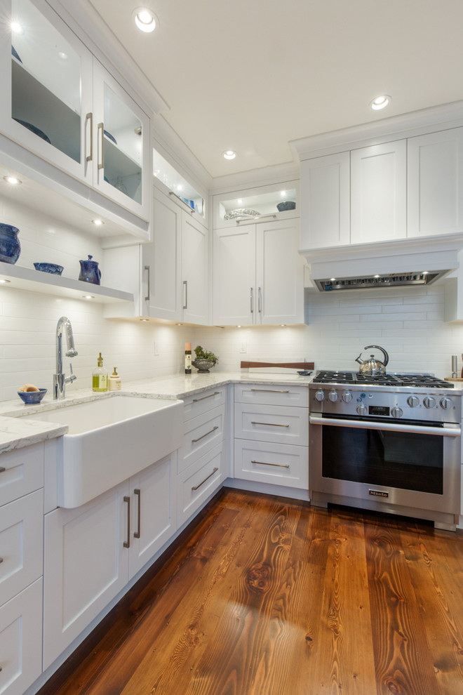 This is an example of a large transitional l-shaped kitchen pantry in Other with a farmhouse sink, shaker cabinets, white cabinets, quartz benchtops, white splashback, subway tile splashback, stainless steel appliances, medium hardwood floors, with island and brown floor.