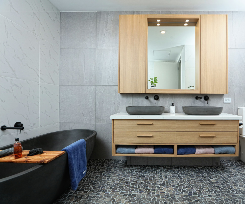 Mid-sized contemporary master bathroom in Melbourne with light wood cabinets, a freestanding tub, porcelain tile, a vessel sink, engineered quartz benchtops, multi-coloured floor, grey walls, mosaic tile floors, white benchtops and flat-panel cabinets.