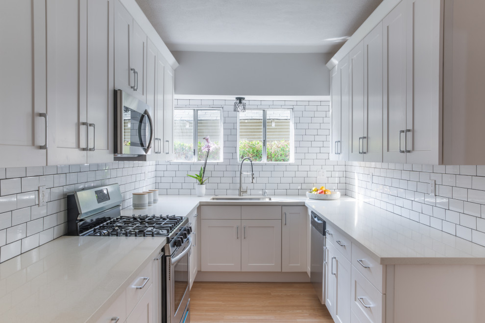 Small contemporary galley kitchen in Los Angeles with an undermount sink, shaker cabinets, white cabinets, solid surface benchtops, white splashback, subway tile splashback, stainless steel appliances, light hardwood floors, no island, brown floor and beige benchtop.