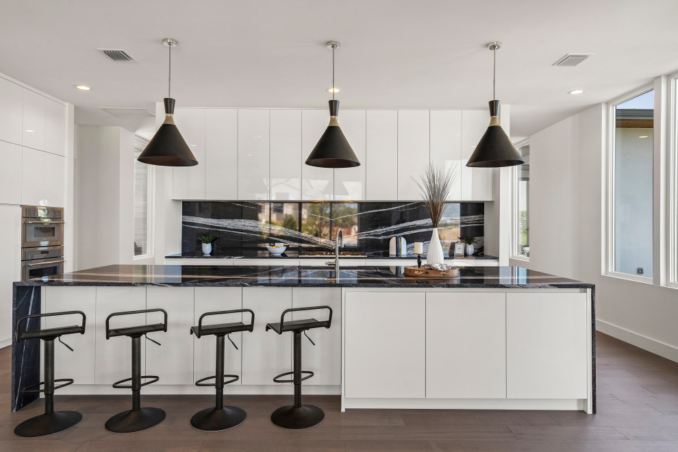 Inspiration for a medium sized contemporary galley kitchen/diner in Austin with a submerged sink, flat-panel cabinets, white cabinets, granite worktops, black splashback, granite splashback, stainless steel appliances, medium hardwood flooring, an island, brown floors, black worktops and all types of ceiling.