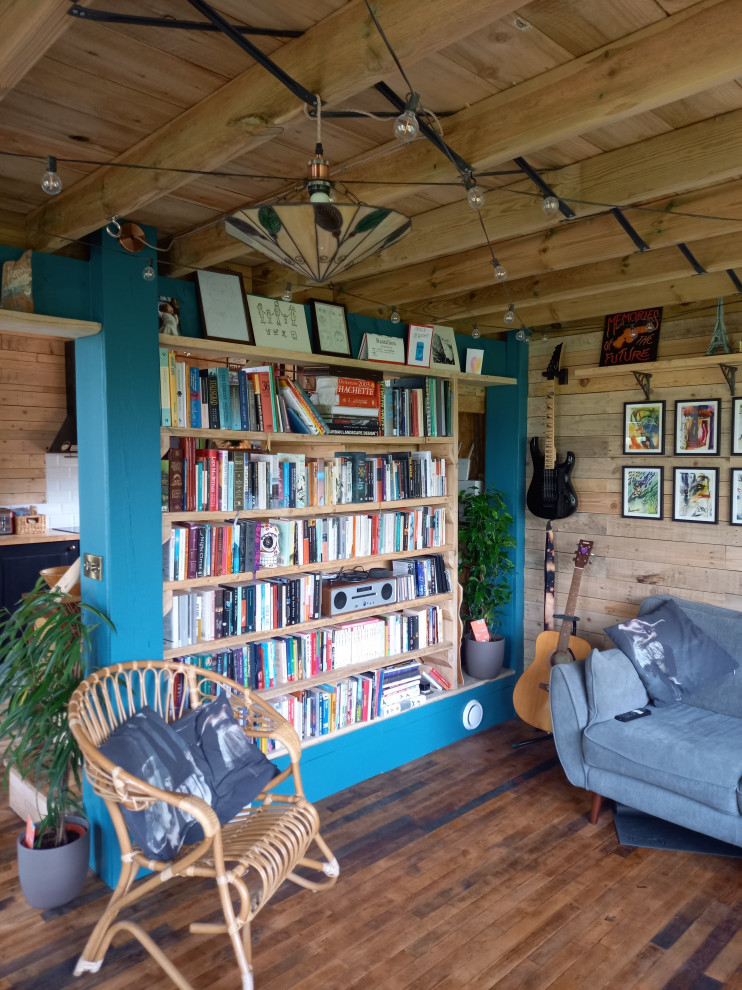 Modelo de biblioteca en casa abierta rural pequeña con paredes marrones, suelo de madera en tonos medios, televisor retractable, suelo marrón, vigas vistas y madera