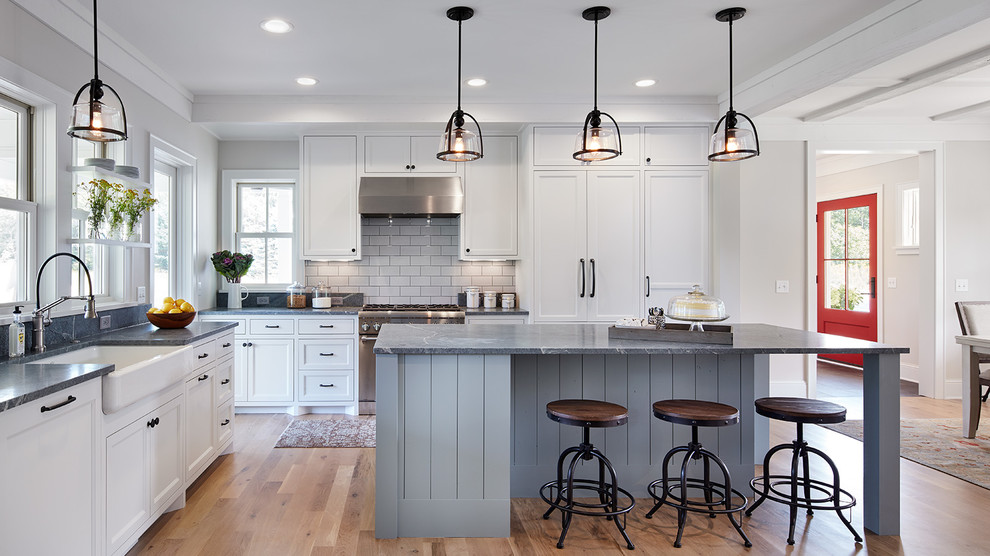 Design ideas for a country l-shaped kitchen in Minneapolis with a farmhouse sink, shaker cabinets, white cabinets, white splashback, subway tile splashback, medium hardwood floors and with island.