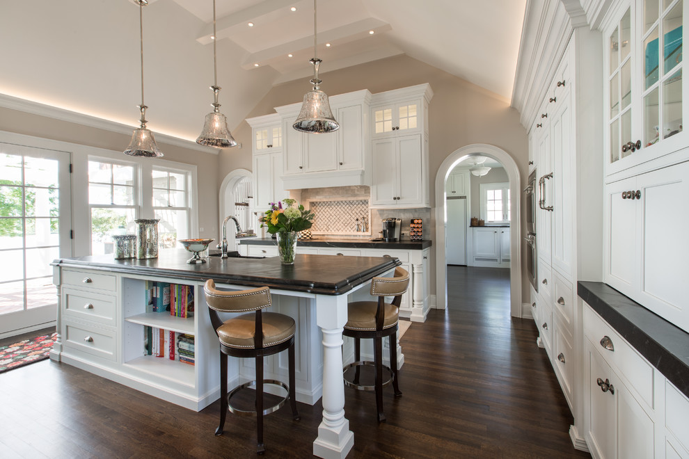 Large u-shaped dark wood floor and brown floor open concept kitchen photo with an island, a farmhouse sink, recessed-panel cabinets, white cabinets, beige backsplash, stainless steel appliances, soapstone countertops and mosaic tile backsplash