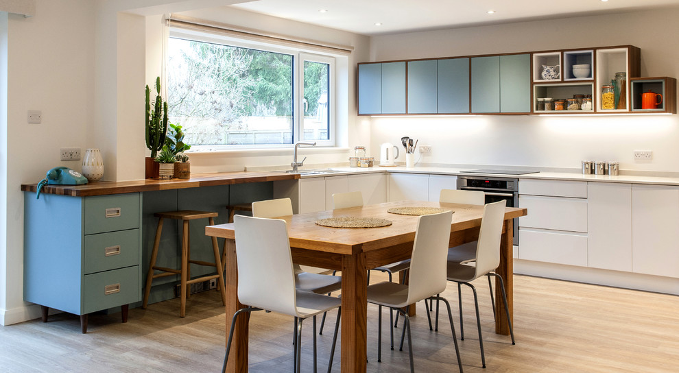 This is an example of a midcentury l-shaped eat-in kitchen in Hampshire with flat-panel cabinets, blue cabinets, wood benchtops, white splashback, stainless steel appliances, light hardwood floors and no island.