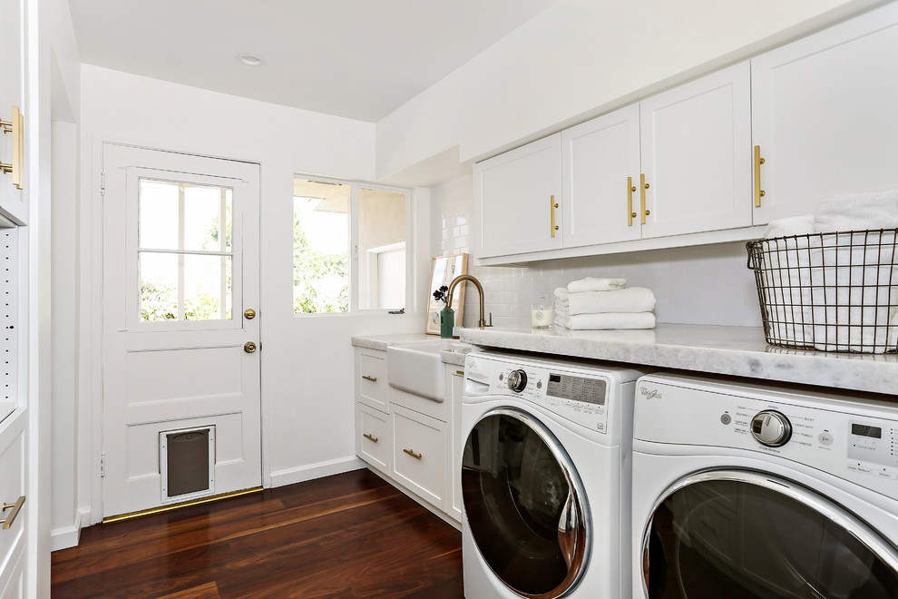 This is an example of a transitional single-wall dedicated laundry room in San Francisco with a farmhouse sink, shaker cabinets, white cabinets, white walls, a side-by-side washer and dryer, brown floor and dark hardwood floors.
