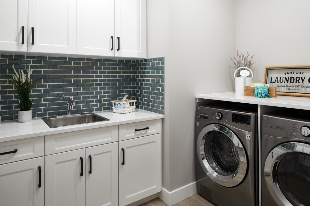 Mid-sized transitional l-shaped utility room in Edmonton with a drop-in sink, shaker cabinets, white cabinets, quartzite benchtops, green splashback, subway tile splashback, grey walls, laminate floors, a side-by-side washer and dryer, brown floor and white benchtop.