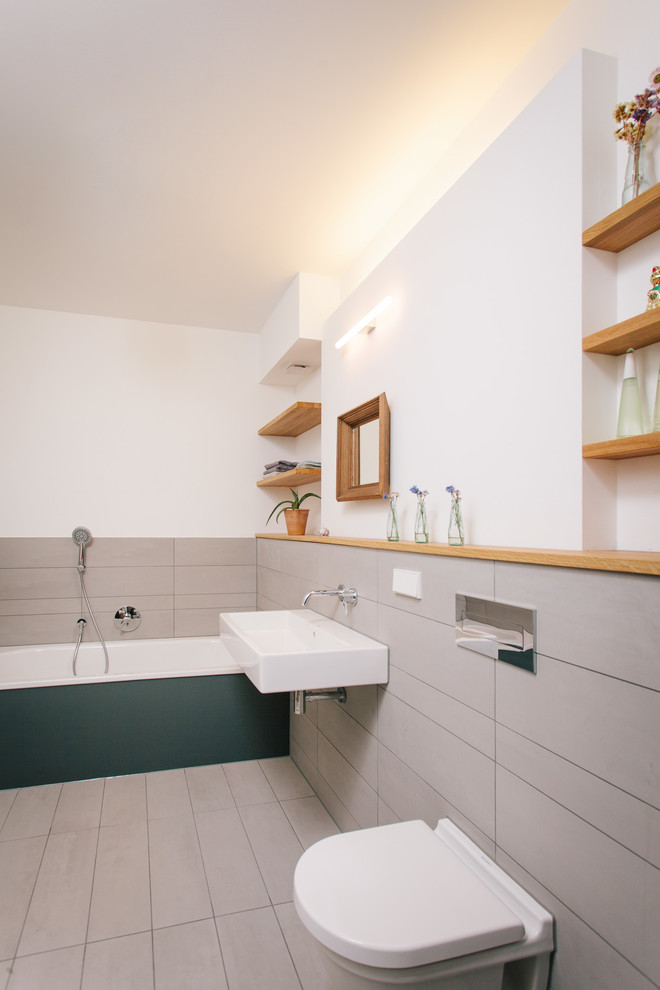 This is an example of a mid-sized contemporary 3/4 bathroom in Cologne with open cabinets, medium wood cabinets, a drop-in tub, a shower/bathtub combo, a wall-mount toilet, gray tile, porcelain tile, white walls, porcelain floors, a wall-mount sink and grey floor.