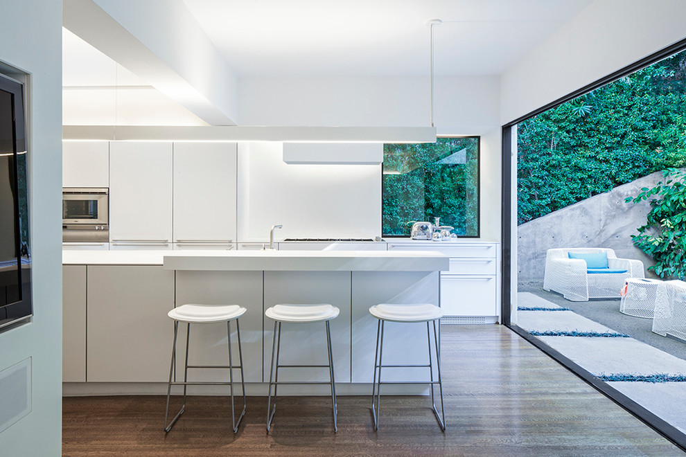 This is an example of a mid-sized contemporary galley eat-in kitchen in Los Angeles with flat-panel cabinets, white cabinets, dark hardwood floors, with island, solid surface benchtops, white splashback and stainless steel appliances.
