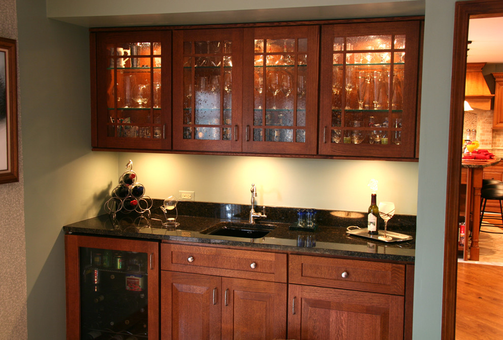 This is an example of a small traditional single-wall wet bar in Chicago with an undermount sink, medium wood cabinets, granite benchtops, medium hardwood floors, recessed-panel cabinets and green splashback.