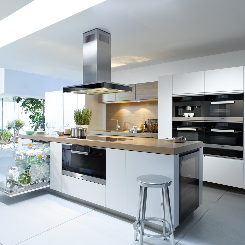 Large contemporary galley open plan kitchen in Oxfordshire with white cabinets, wood benchtops, brown splashback, black appliances, ceramic floors and with island.