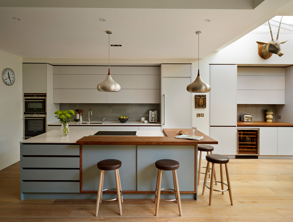 This is an example of a large contemporary single-wall open plan kitchen in London with flat-panel cabinets, blue cabinets, solid surface benchtops, grey splashback, glass tile splashback, stainless steel appliances, light hardwood floors, with island and white benchtop.