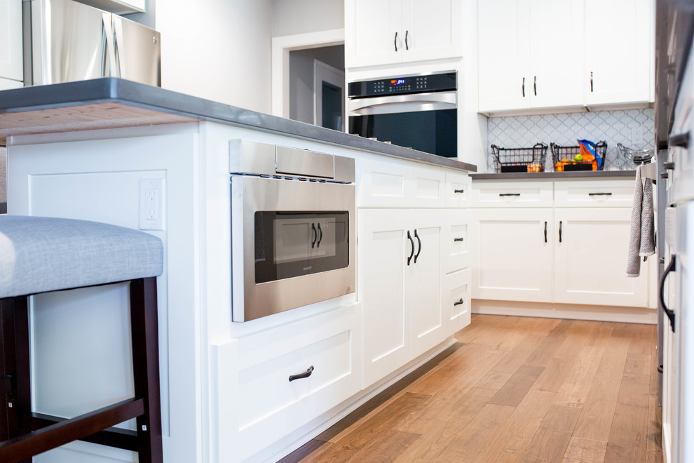 Example of a mid-sized transitional light wood floor eat-in kitchen design in Sacramento with a farmhouse sink, shaker cabinets, white cabinets, white backsplash, glass tile backsplash, stainless steel appliances and an island