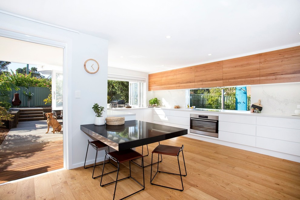 Inspiration for a contemporary u-shaped eat-in kitchen in Wollongong with flat-panel cabinets, white cabinets, white splashback, stone slab splashback, stainless steel appliances, medium hardwood floors, brown floor and white benchtop.