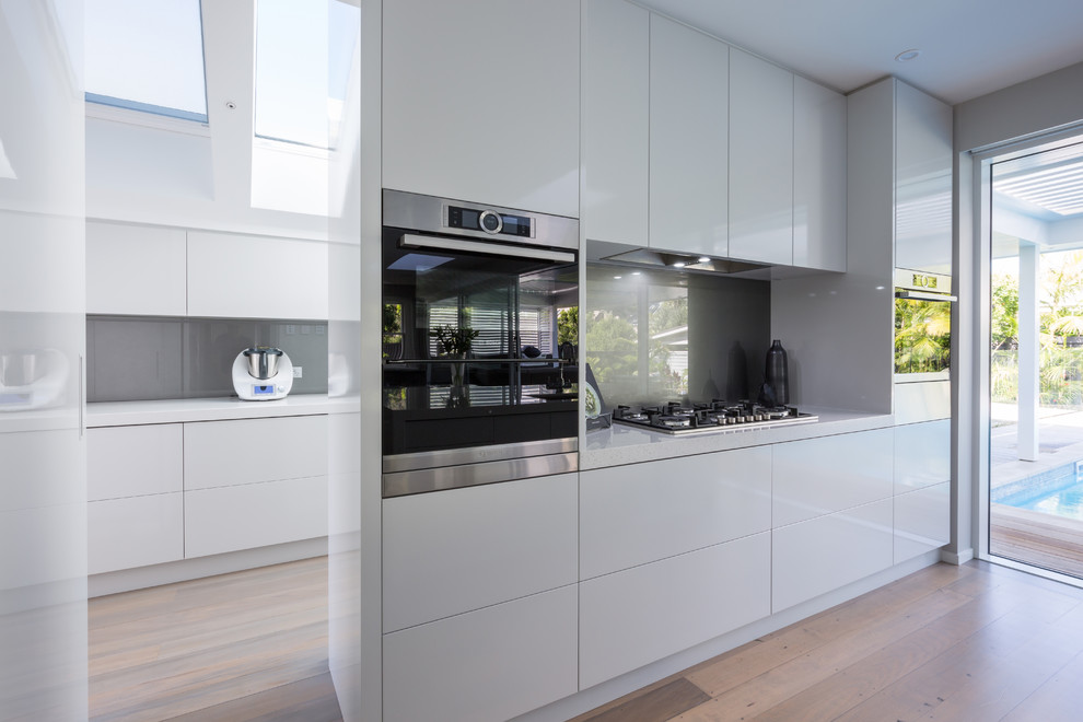 Large contemporary l-shaped eat-in kitchen in Auckland with an undermount sink, flat-panel cabinets, grey cabinets, quartz benchtops, grey splashback, glass sheet splashback, stainless steel appliances, light hardwood floors, with island and beige floor.
