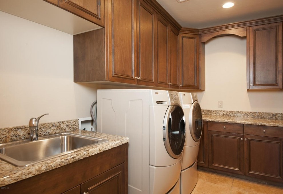 Photo of a traditional l-shaped dedicated laundry room in Phoenix with a drop-in sink, recessed-panel cabinets, medium wood cabinets, granite benchtops, beige walls, travertine floors and a side-by-side washer and dryer.