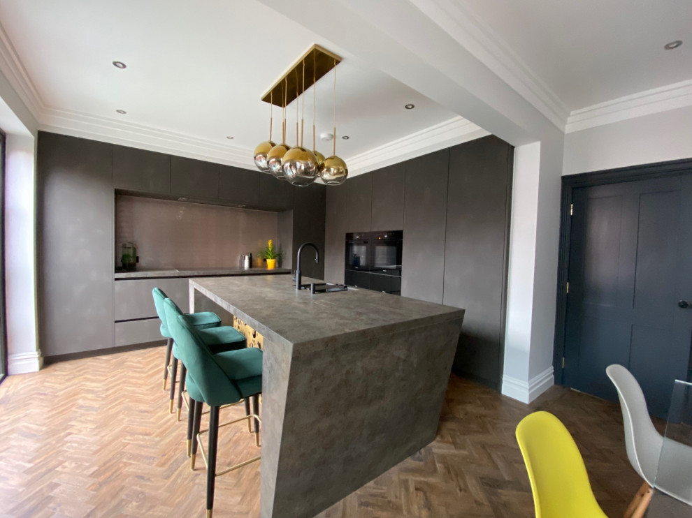 This is an example of a mid-sized contemporary l-shaped eat-in kitchen in Manchester with an undermount sink, solid surface benchtops, metallic splashback, glass sheet splashback, black appliances, medium hardwood floors, with island, brown floor and grey benchtop.