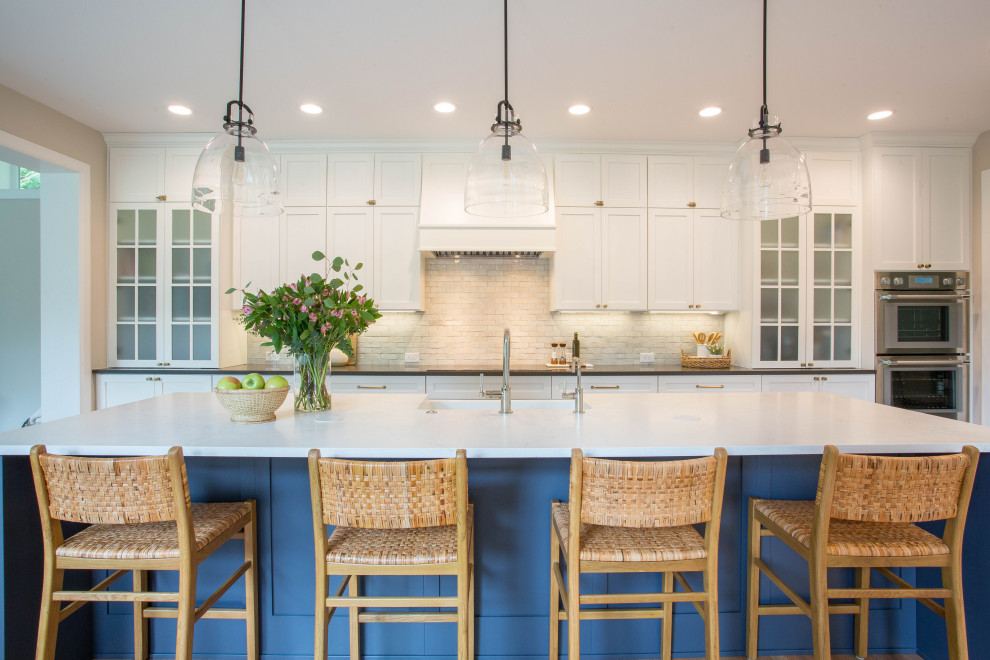 This is an example of a large country single-wall open plan kitchen in Seattle with a farmhouse sink, shaker cabinets, white cabinets, quartz benchtops, grey splashback, cement tile splashback, stainless steel appliances, light hardwood floors, with island, brown floor and white benchtop.