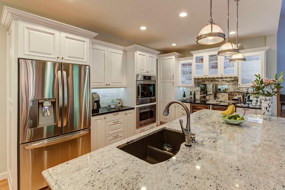 Mid-sized transitional galley light wood floor eat-in kitchen photo in Raleigh with a double-bowl sink, raised-panel cabinets, white cabinets, granite countertops, white backsplash, stone tile backsplash, stainless steel appliances and an island