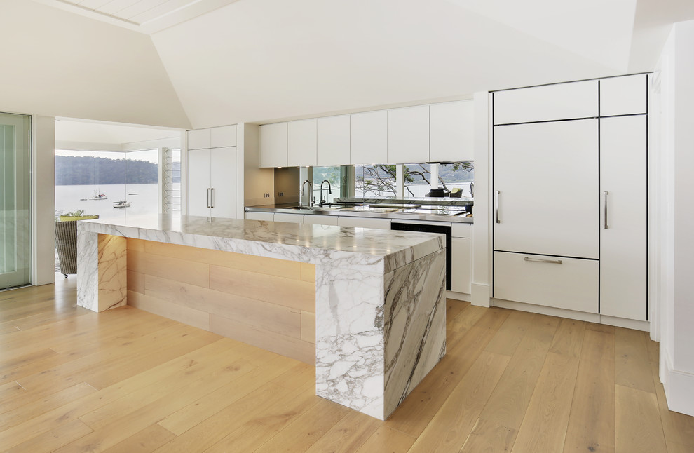 This is an example of a large modern single-wall open plan kitchen in Sydney with an integrated sink, flat-panel cabinets, beige cabinets, marble benchtops, metallic splashback, mirror splashback, black appliances, light hardwood floors and with island.