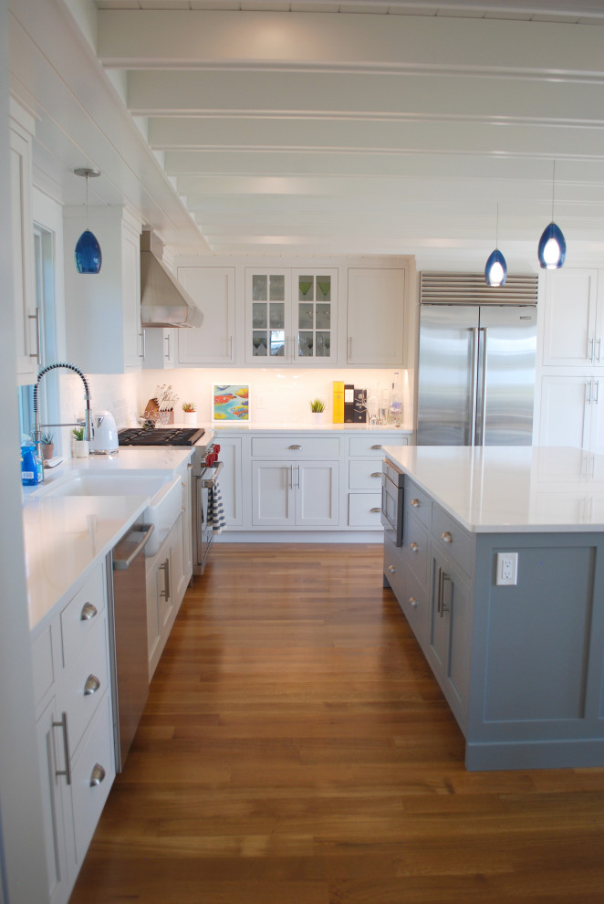 Design ideas for a large beach style u-shaped eat-in kitchen in Providence with a farmhouse sink, shaker cabinets, white cabinets, granite benchtops, white splashback, subway tile splashback, stainless steel appliances, light hardwood floors, with island, white benchtop and exposed beam.