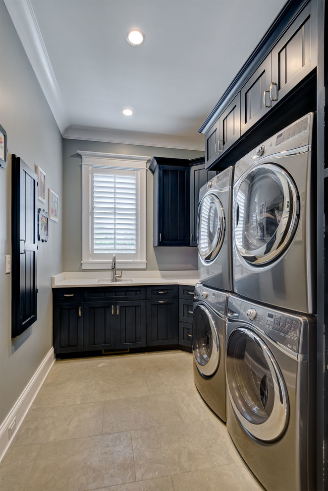 Inspiration for a large transitional l-shaped dedicated laundry room in Chicago with an undermount sink, recessed-panel cabinets, black cabinets, quartz benchtops, grey walls, travertine floors, a stacked washer and dryer, brown floor and white benchtop.