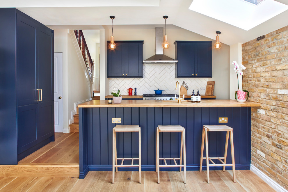 Photo of a transitional u-shaped kitchen in London with an undermount sink, shaker cabinets, blue cabinets, wood benchtops, white splashback, panelled appliances, medium hardwood floors, a peninsula, brown floor and brown benchtop.