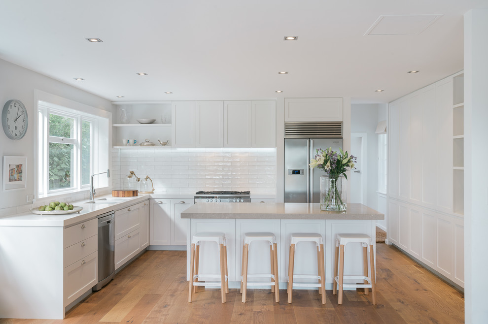Design ideas for a transitional l-shaped kitchen in Christchurch with a double-bowl sink, shaker cabinets, white cabinets, white splashback, subway tile splashback, stainless steel appliances, medium hardwood floors and with island.