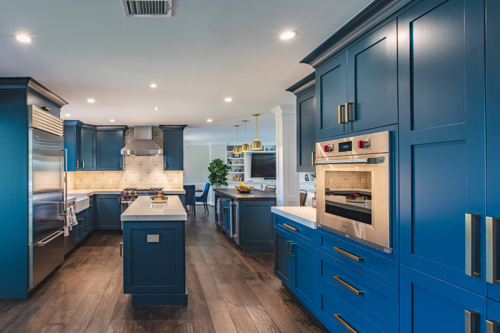Mid-sized transitional u-shaped eat-in kitchen in DC Metro with a farmhouse sink, recessed-panel cabinets, blue cabinets, quartz benchtops, multi-coloured splashback, porcelain splashback, stainless steel appliances, medium hardwood floors, multiple islands, brown floor and grey benchtop.