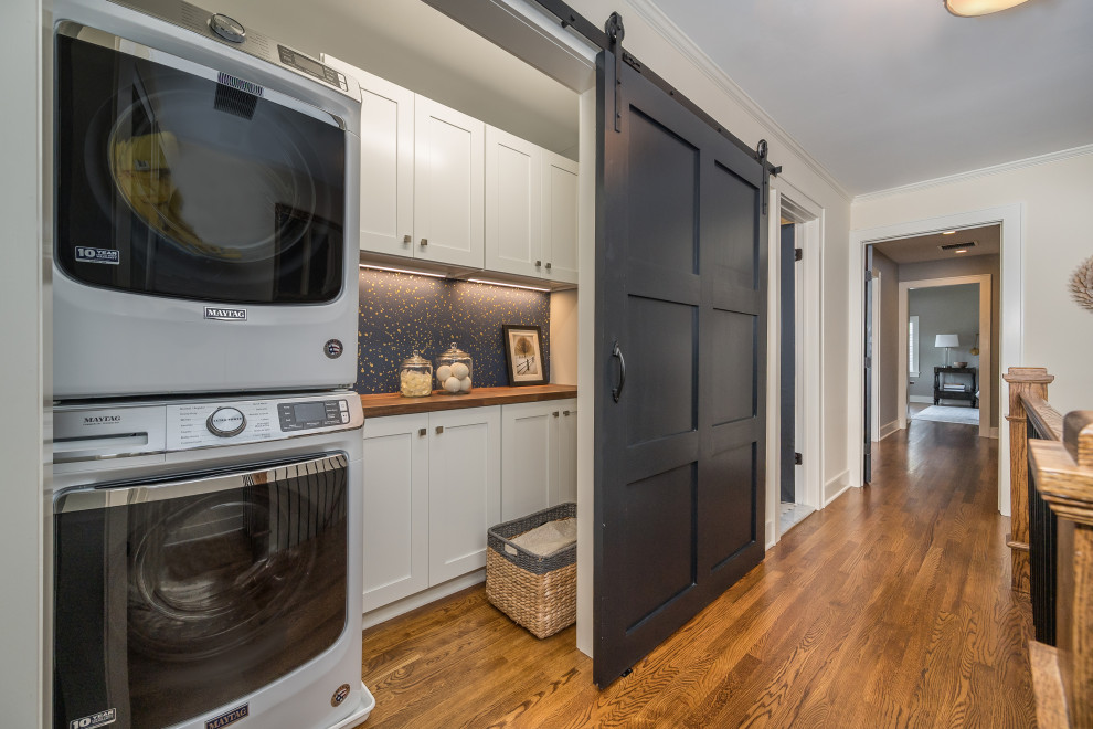 Inspiration for a mid-sized transitional single-wall laundry cupboard in Newark with recessed-panel cabinets, white cabinets, wood benchtops, blue splashback, blue walls, light hardwood floors, a stacked washer and dryer, brown floor and brown benchtop.