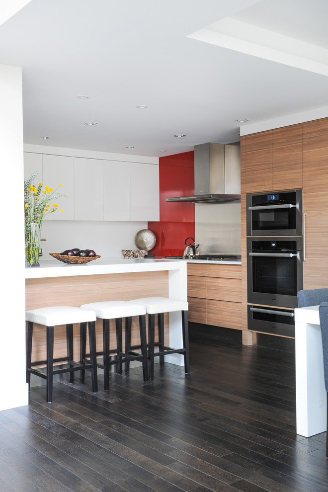 Photo of a mid-sized contemporary l-shaped eat-in kitchen in Vancouver with a drop-in sink, flat-panel cabinets, medium wood cabinets, quartz benchtops, red splashback, glass sheet splashback, stainless steel appliances, dark hardwood floors and a peninsula.