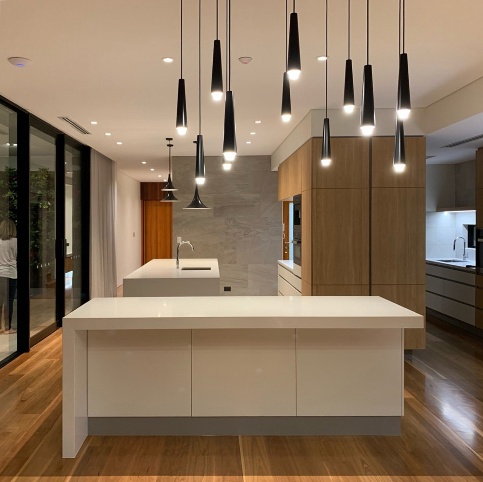 Example of a large trendy l-shaped medium tone wood floor and brown floor kitchen pantry design in Perth with an undermount sink, flat-panel cabinets, white cabinets, limestone countertops, two islands, white countertops and stainless steel appliances