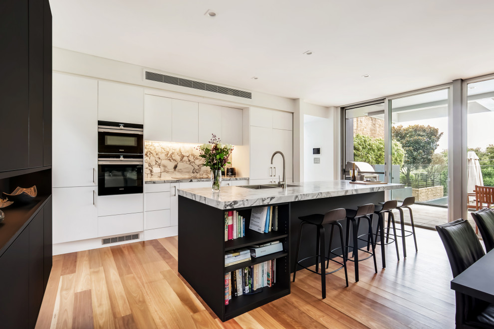 Inspiration for a mid-sized contemporary galley open plan kitchen in Sydney with a drop-in sink, flat-panel cabinets, marble benchtops, grey splashback, stone slab splashback, stainless steel appliances, medium hardwood floors, with island and grey benchtop.