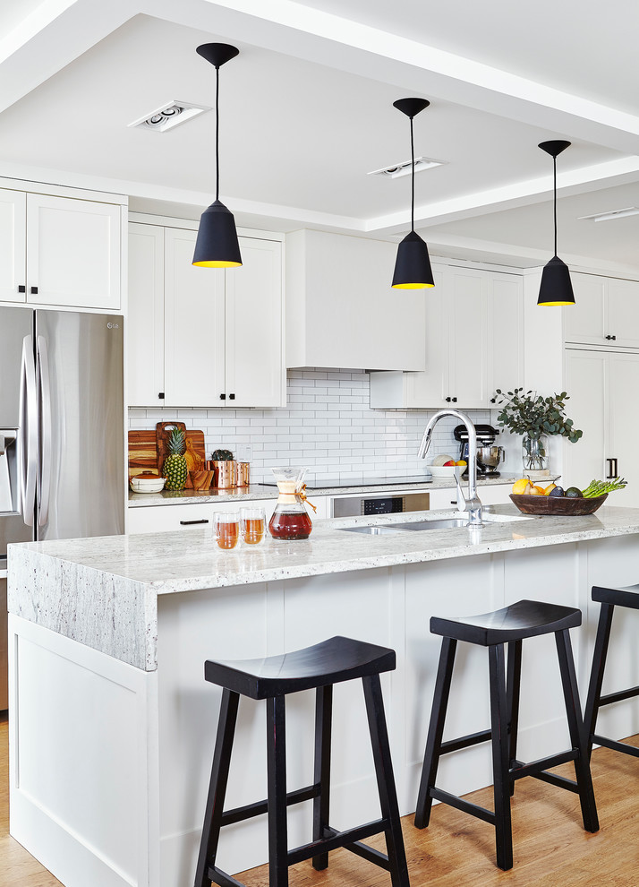 Design ideas for a mid-sized transitional galley kitchen in Toronto with an undermount sink, shaker cabinets, white cabinets, granite benchtops, white splashback, ceramic splashback, stainless steel appliances, with island, medium hardwood floors and brown floor.