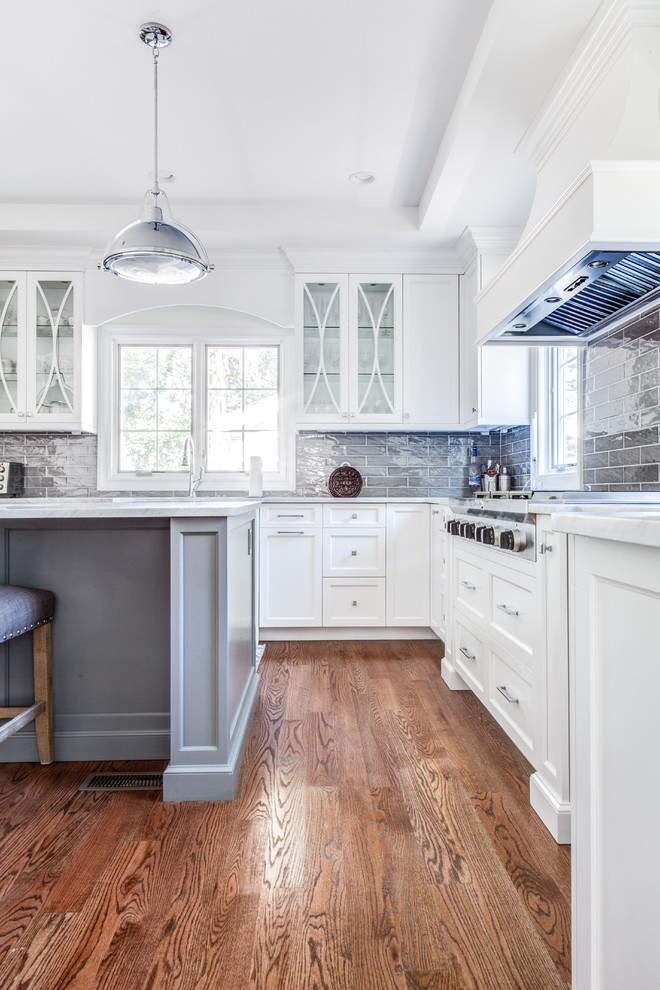 Design ideas for a mid-sized transitional u-shaped open plan kitchen in New York with a farmhouse sink, shaker cabinets, white cabinets, quartz benchtops, grey splashback, glass tile splashback, panelled appliances, light hardwood floors, with island and brown floor.