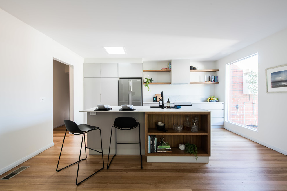 This is an example of a mid-sized contemporary galley kitchen in Melbourne with medium hardwood floors, with island, white benchtop, an undermount sink, flat-panel cabinets, white cabinets, white splashback, stainless steel appliances and brown floor.