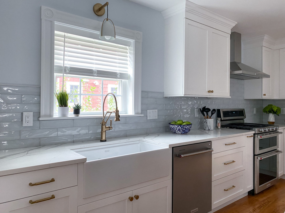 Photo of a traditional kitchen/diner in Boston with a belfast sink, shaker cabinets, white cabinets, engineered stone countertops, blue splashback, glass tiled splashback, stainless steel appliances, medium hardwood flooring, an island, brown floors and white worktops.