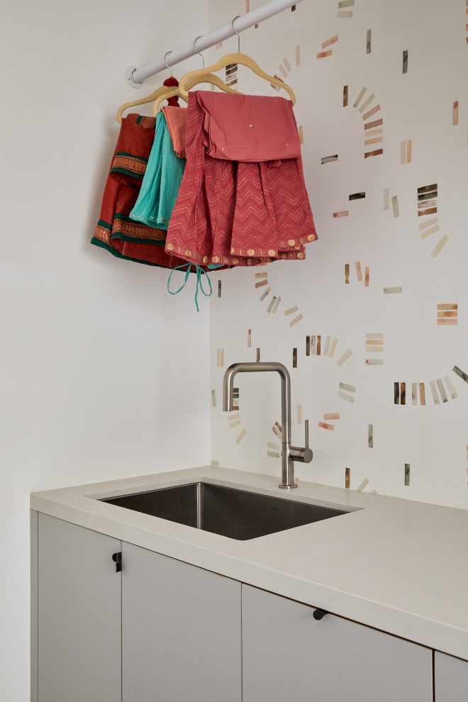 This is an example of a mid-sized modern galley dedicated laundry room in San Francisco with an undermount sink, flat-panel cabinets, grey cabinets, quartz benchtops, white walls, porcelain floors, a side-by-side washer and dryer, grey floor, grey benchtop and wallpaper.