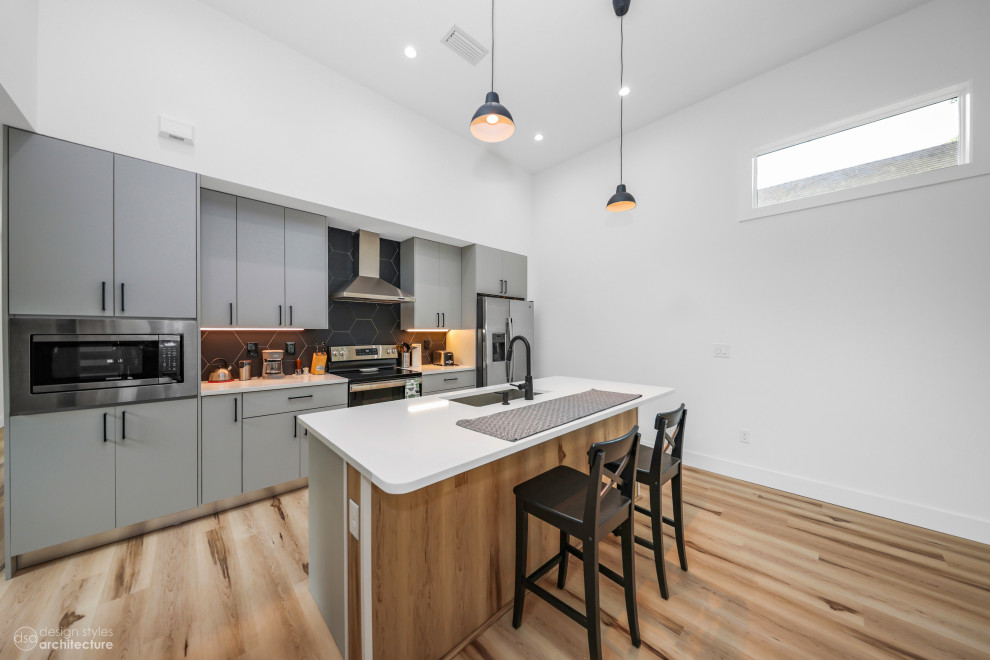 Inspiration for a mid-sized contemporary single-wall open plan kitchen in Tampa with a drop-in sink, flat-panel cabinets, grey cabinets, quartz benchtops, black splashback, cement tile splashback, stainless steel appliances, light hardwood floors, with island, brown floor and white benchtop.