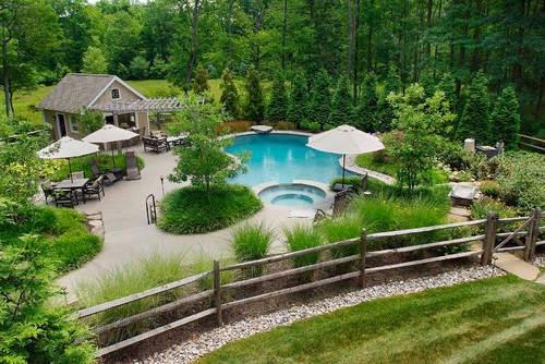 An enclosed backyard viewed from above showing the pool complex, including several patio areas, a garden, and a pool house. The area is enclosed by a large, sturdy mortised fence.