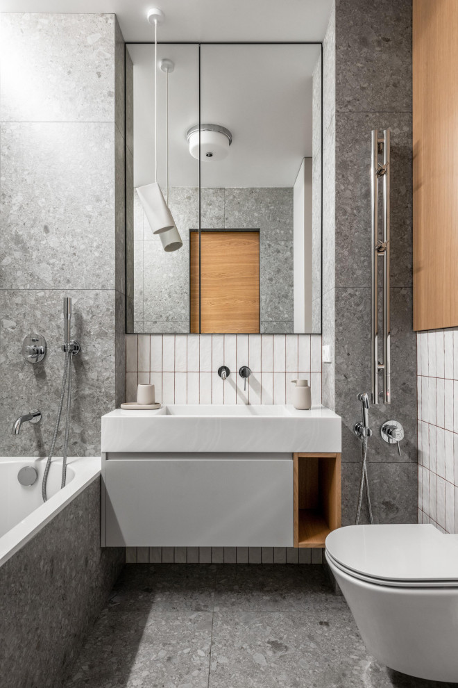 Bild på ett funkis badrum, med släta luckor, vita skåp, ett badkar i en alkov, en dusch/badkar-kombination, en vägghängd toalettstol, vit kakel, ett konsol handfat och grått golv