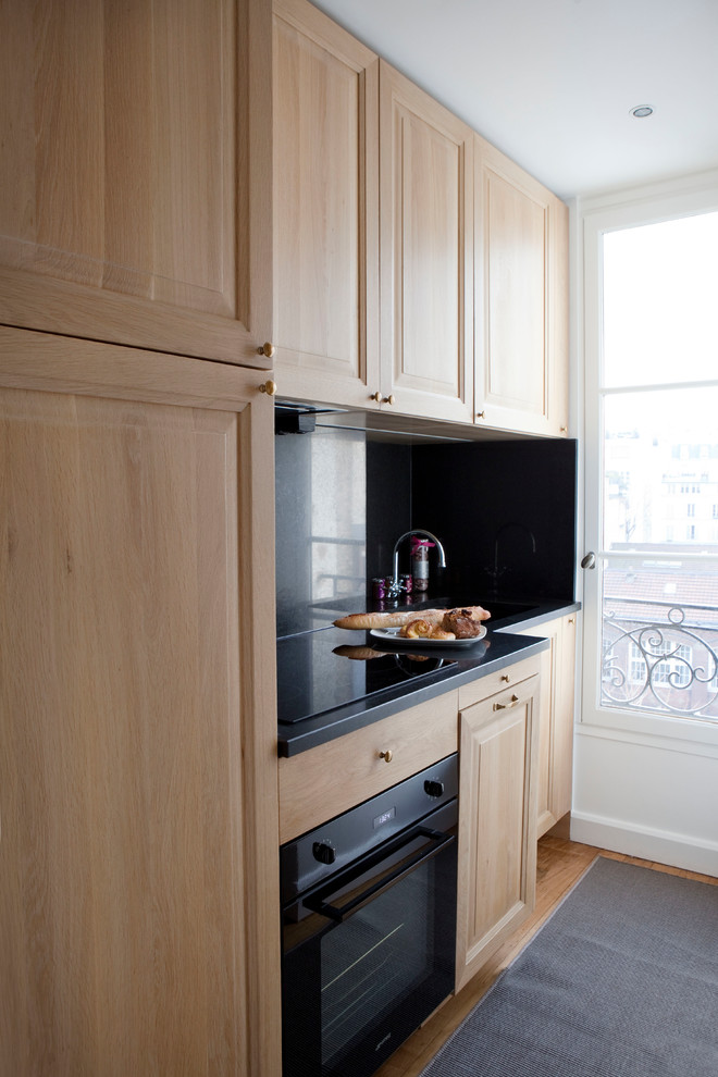 Inspiration for a small transitional single-wall separate kitchen in Paris with an integrated sink, beaded inset cabinets, light wood cabinets, granite benchtops, black splashback, limestone splashback, black appliances, light hardwood floors, no island, brown floor and black benchtop.