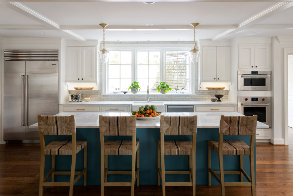 Photo of an expansive country open plan kitchen in Philadelphia with a farmhouse sink, raised-panel cabinets, white cabinets, white splashback, porcelain splashback, stainless steel appliances, medium hardwood floors, with island, brown floor, coffered, marble benchtops and white benchtop.