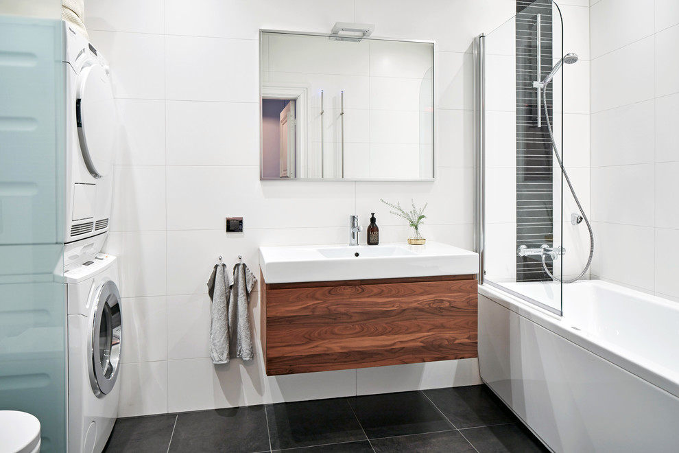 Mid-sized contemporary bathroom in Gothenburg with flat-panel cabinets, medium wood cabinets, a drop-in tub, a shower/bathtub combo, white tile, an integrated sink, porcelain tile, white walls, limestone floors and a laundry.