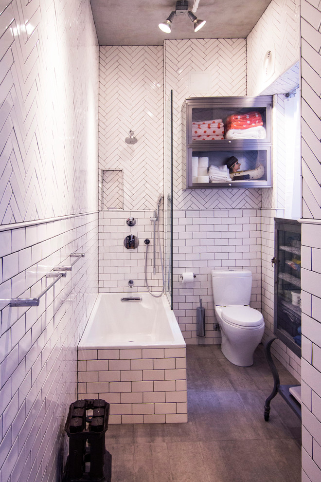 Photo of a small industrial master bathroom in New York with a wall-mount sink, glass-front cabinets, grey cabinets, a drop-in tub, a corner shower, white tile, white walls, porcelain floors, subway tile, brown floor and a one-piece toilet.