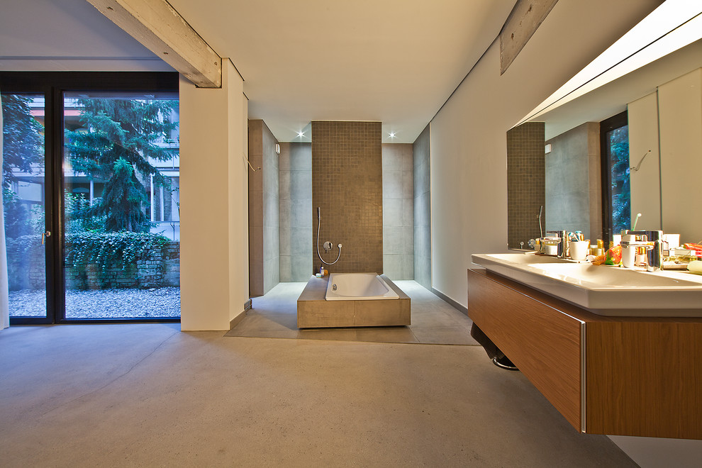 Expansive industrial bathroom in Stuttgart with flat-panel cabinets, medium wood cabinets, a drop-in tub, gray tile, white walls, concrete floors and an integrated sink.