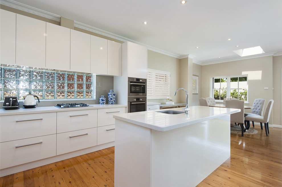 This is an example of a transitional galley eat-in kitchen in Sydney with an integrated sink, white cabinets, limestone benchtops, grey splashback, glass sheet splashback, stainless steel appliances and with island.
