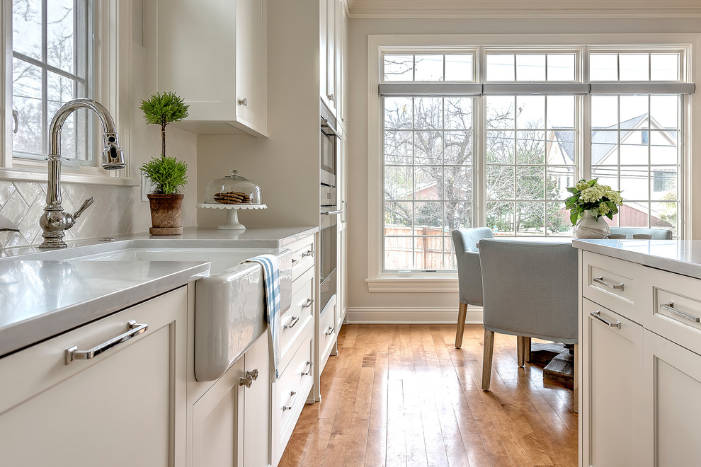 Photo of a mid-sized country l-shaped eat-in kitchen in Chicago with a farmhouse sink, shaker cabinets, white cabinets, quartz benchtops, white splashback, ceramic splashback, panelled appliances, light hardwood floors and with island.