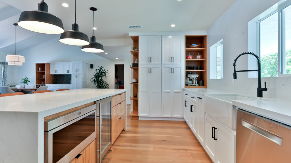 This is an example of a large contemporary open plan kitchen in Los Angeles with a farmhouse sink, shaker cabinets, yellow cabinets, quartz benchtops, white splashback, engineered quartz splashback, stainless steel appliances, medium hardwood floors, with island, brown floor, white benchtop and vaulted.