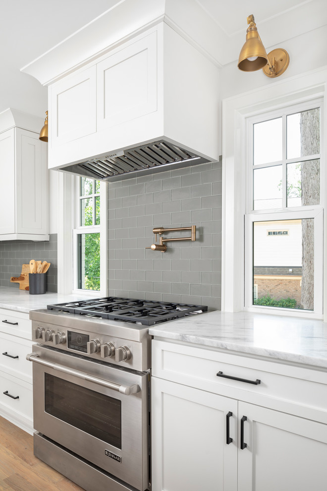 Example of a mid-sized transitional l-shaped medium tone wood floor eat-in kitchen design in Charlotte with a farmhouse sink, white cabinets, marble countertops, gray backsplash, glass tile backsplash, stainless steel appliances, an island and white countertops