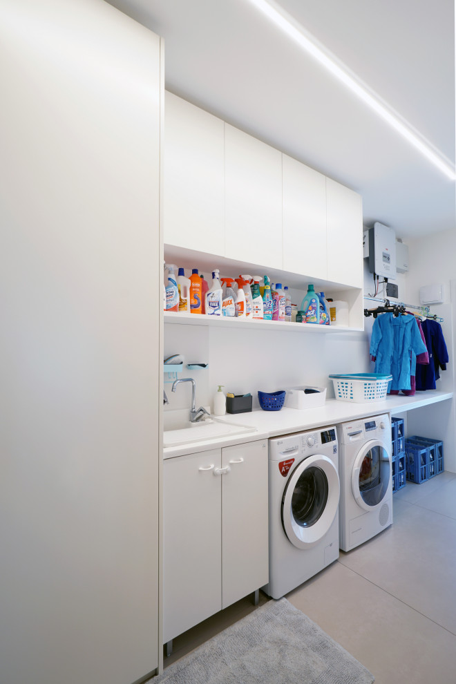 Photo of a small contemporary single-wall utility room in Venice with a drop-in sink, flat-panel cabinets, white cabinets, solid surface benchtops, white splashback, porcelain splashback, white walls, porcelain floors, a side-by-side washer and dryer, beige floor and white benchtop.